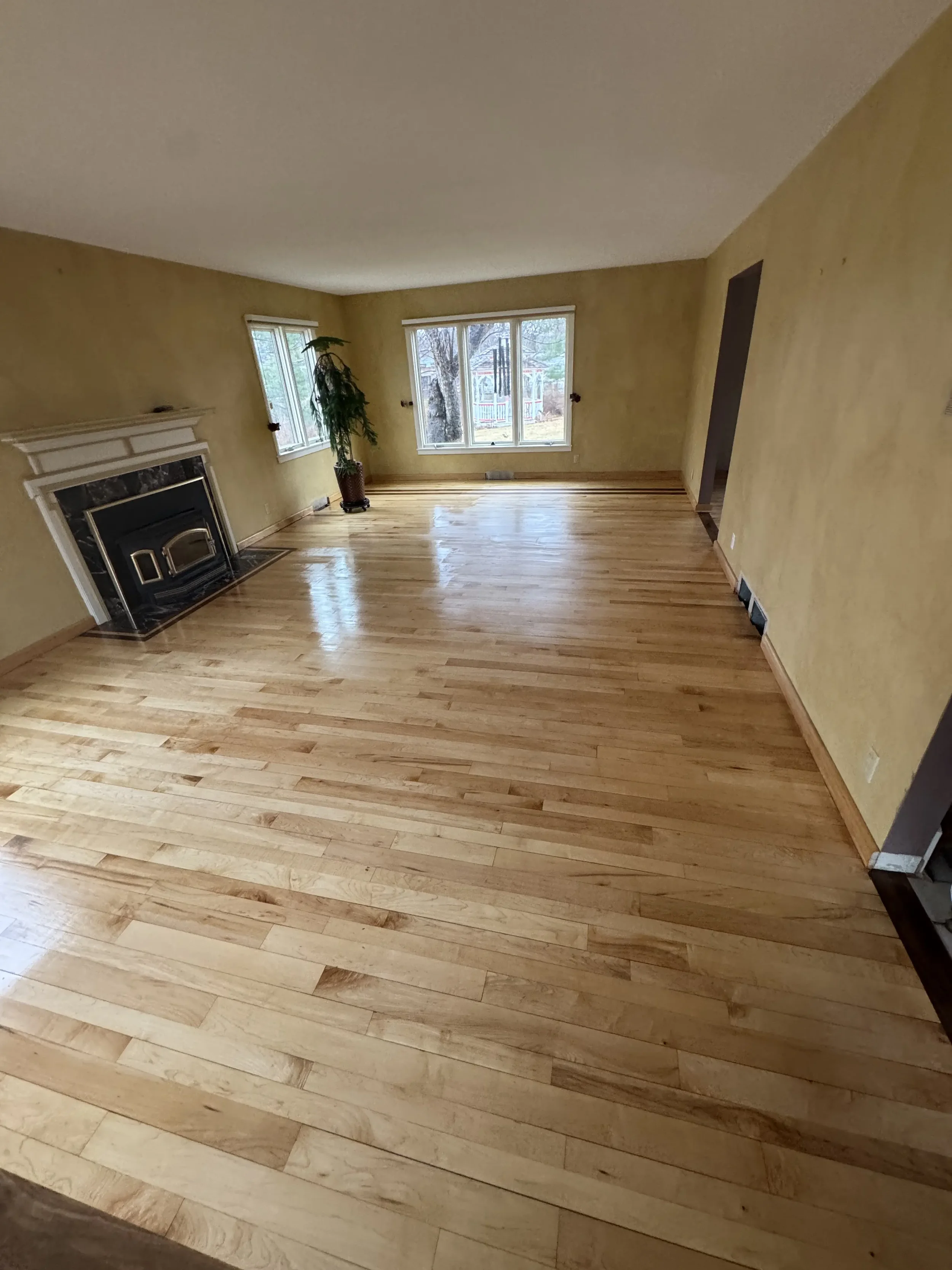 Empty living room with polished wooden floors, yellow walls, a black fireplace, and large windows showing a backyard with trees and a playground.