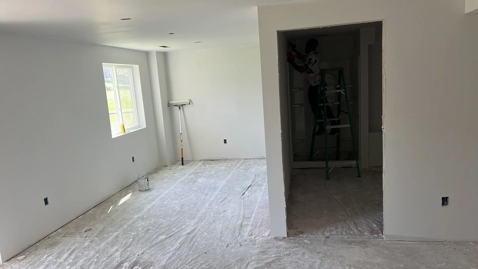 Interior of a house under construction with white painted walls, a window letting in natural light, and construction tools such as a stepladder and a paint roller in the room.