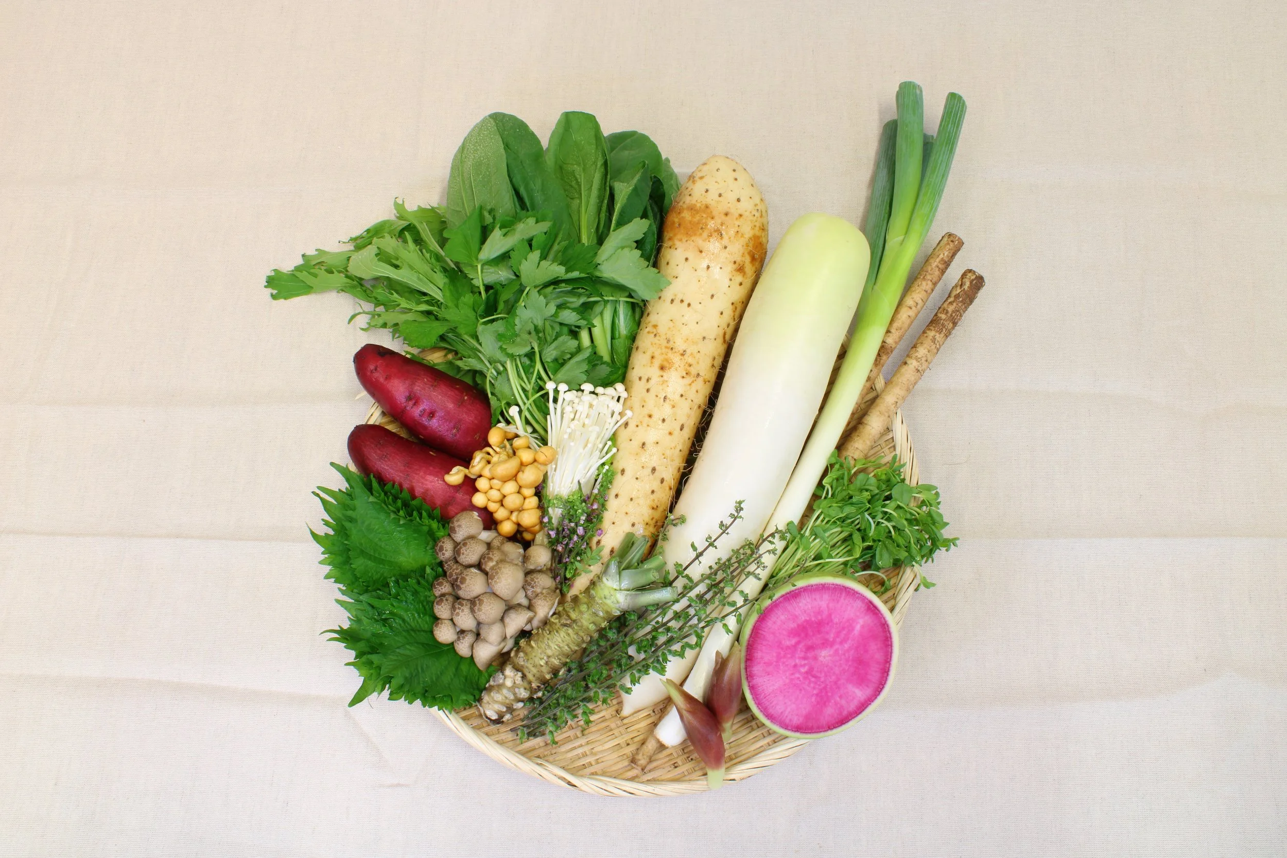 Assorted fresh vegetables including radish, green leafy herbs, parsnip, daikon radish, green onions, and root vegetables arranged on a woven basket.