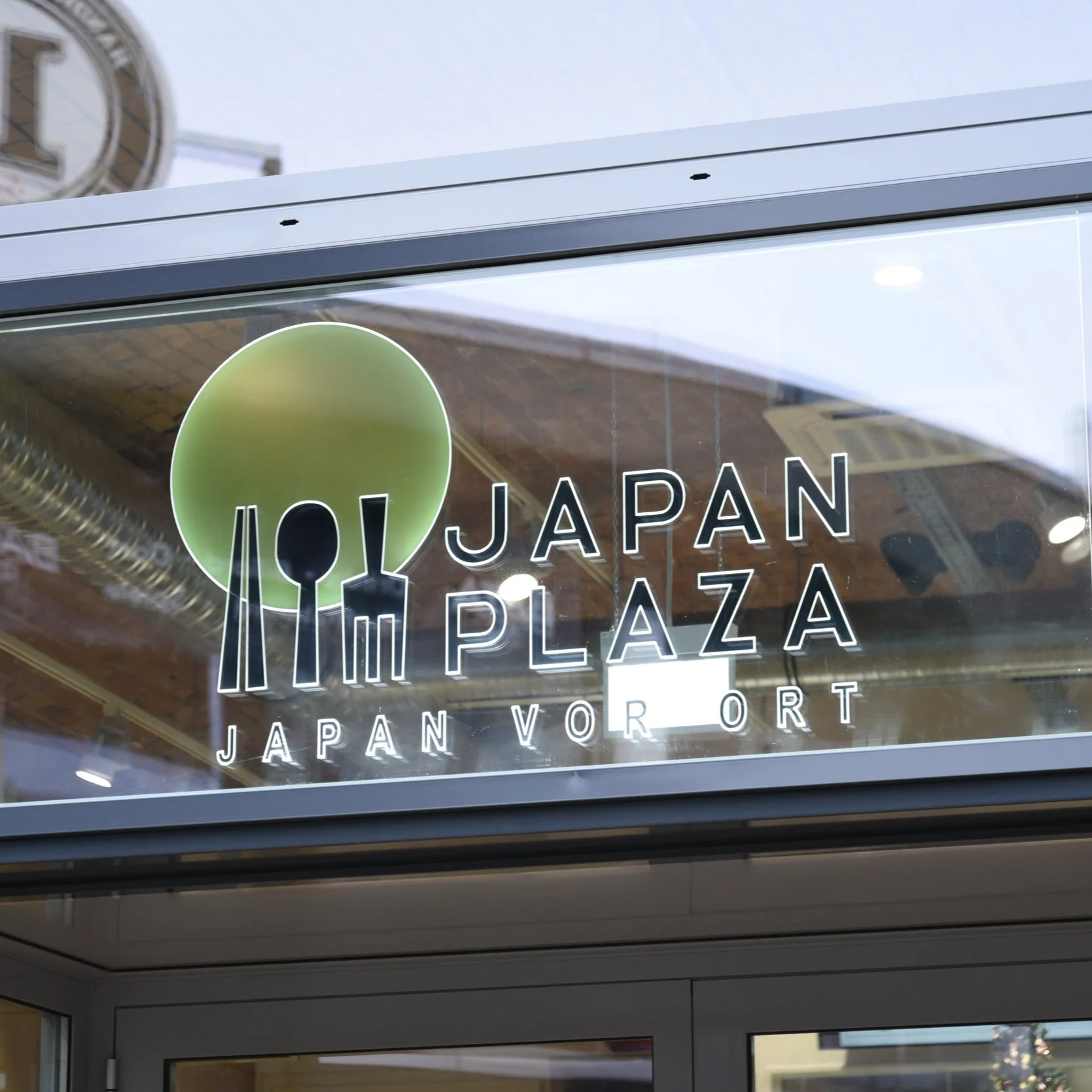 Sign on glass door displaying 'JAPAN PLAZA' with green logo and Japanese text, reflecting buildings outside