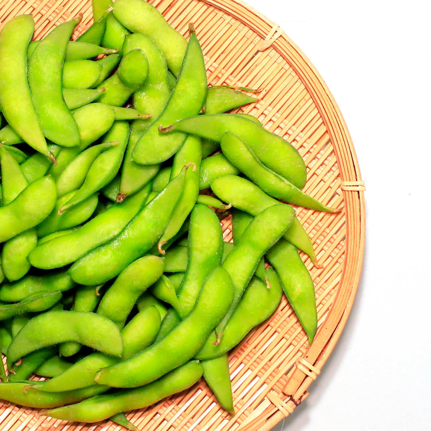 Green edamame beans in a woven basket on a white background.