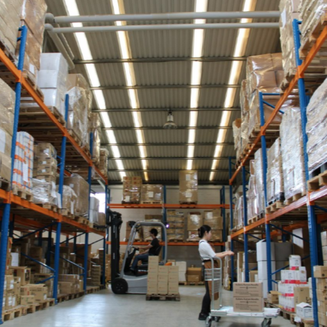 Warehouse with shelves stocked with boxes, a worker operating a forklift, and another worker stacking boxes.