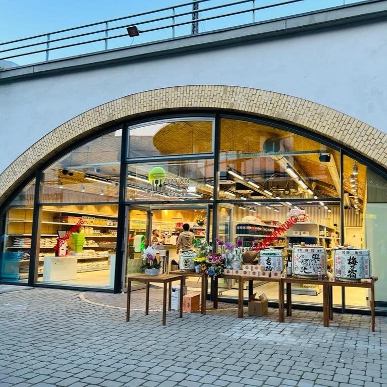 Exterior of a store with large glass windows displaying shelves of products and a few customers inside. Outside, there are tables with flower arrangements and large boxes on display.