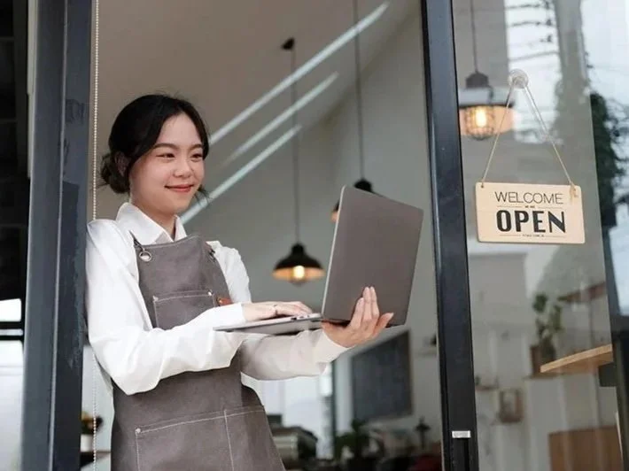A young woman in an apron holding a laptop and smiling at a cafe entrance with a sign that reads 'Welcome Open'.