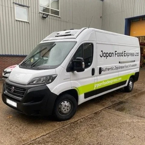 White delivery van with 'Japan Food Express' branding, parked outside a warehouse, with a large blue garage door.