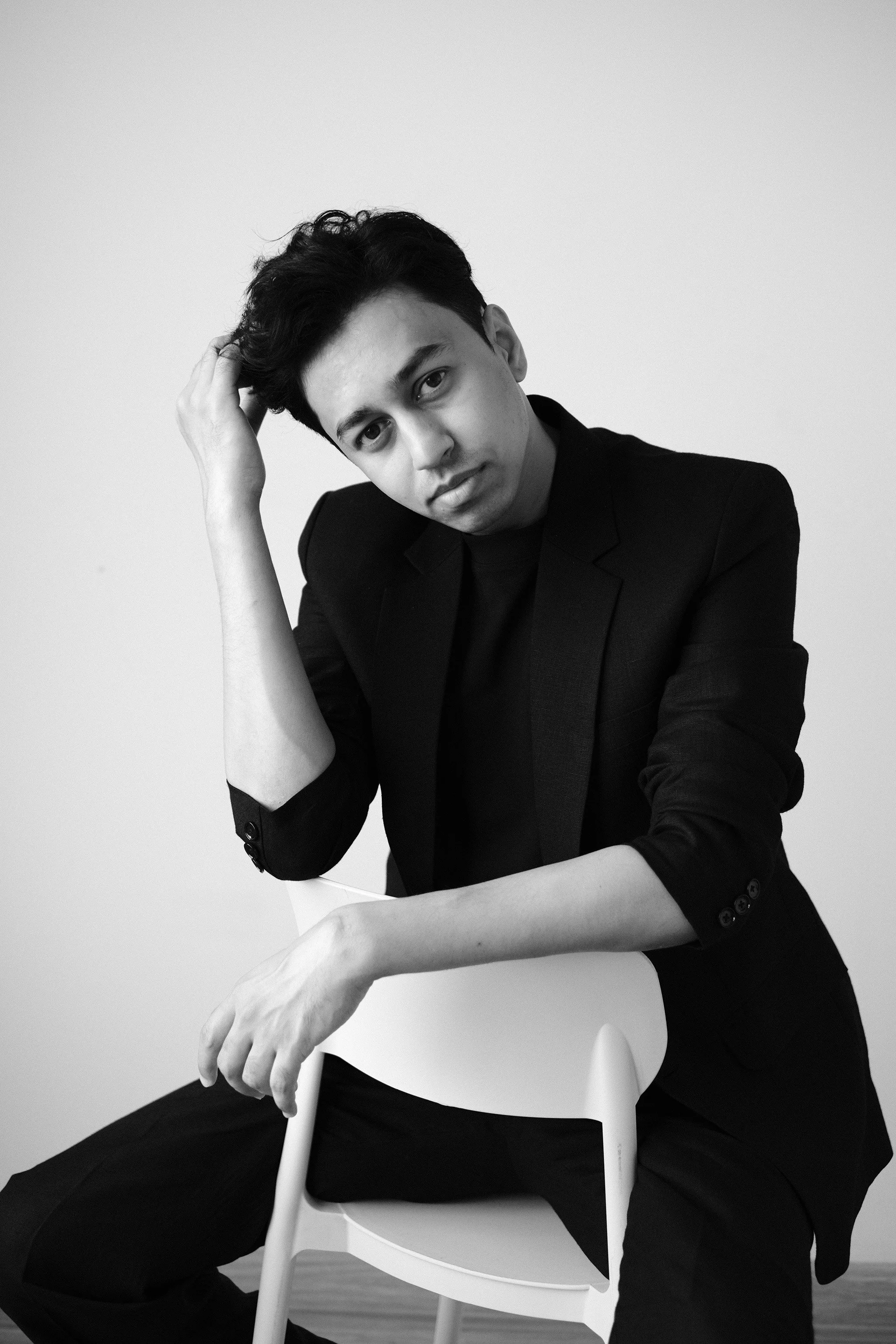 A man with dark hair and light skin, wearing a black blazer, sitting on a white chair, resting his head on his left hand, looking into the camera, black and white photo.