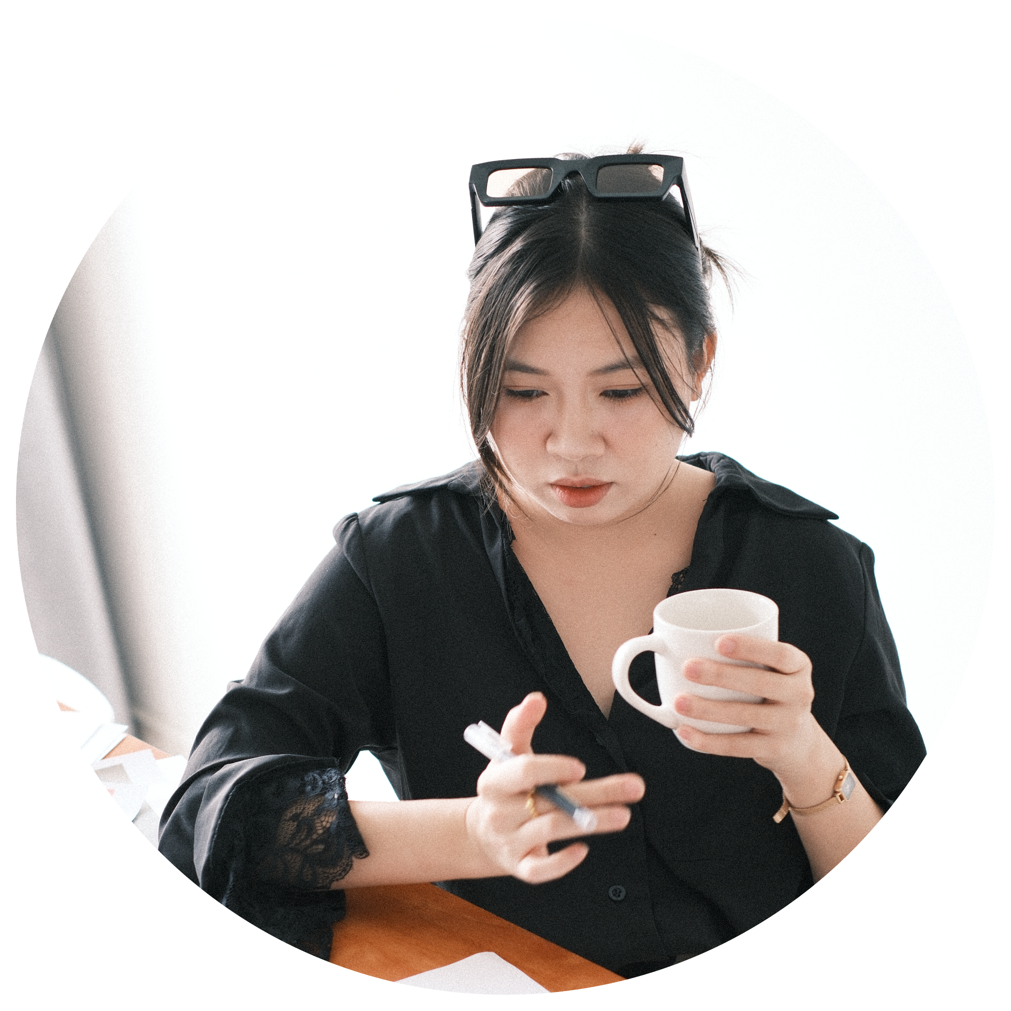 A woman with dark hair wearing glasses resting on her head, looking down at her phone while holding a white mug, with papers on a wooden table.