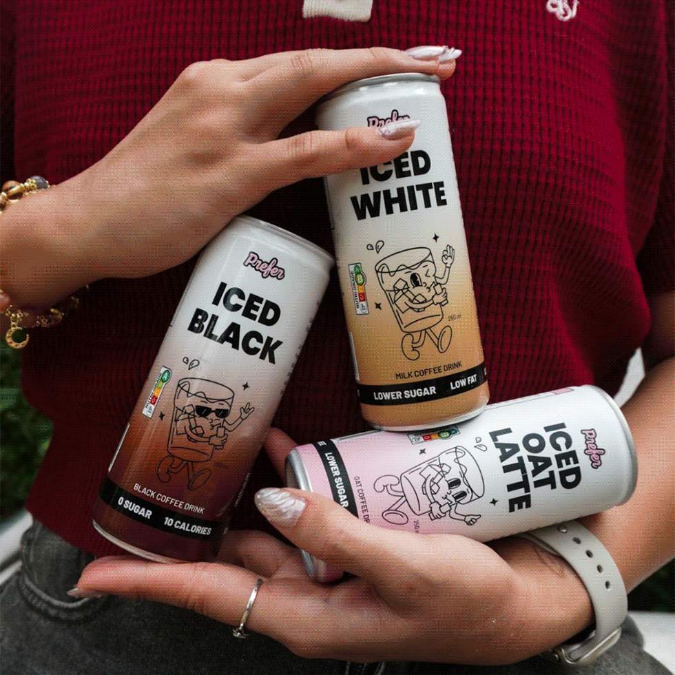 Three cans of flavored coffee drinks held by a person wearing a red shirt and a white watch, with one can labeled "Iced Black," another "Iced White," and the third "Iced Oat Latte."