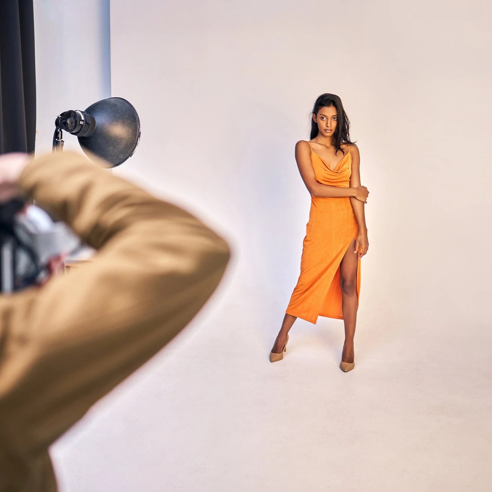 A woman in an orange dress posing for a photoshoot against a white background. There is a professional studio light and a person's arm in the foreground.
