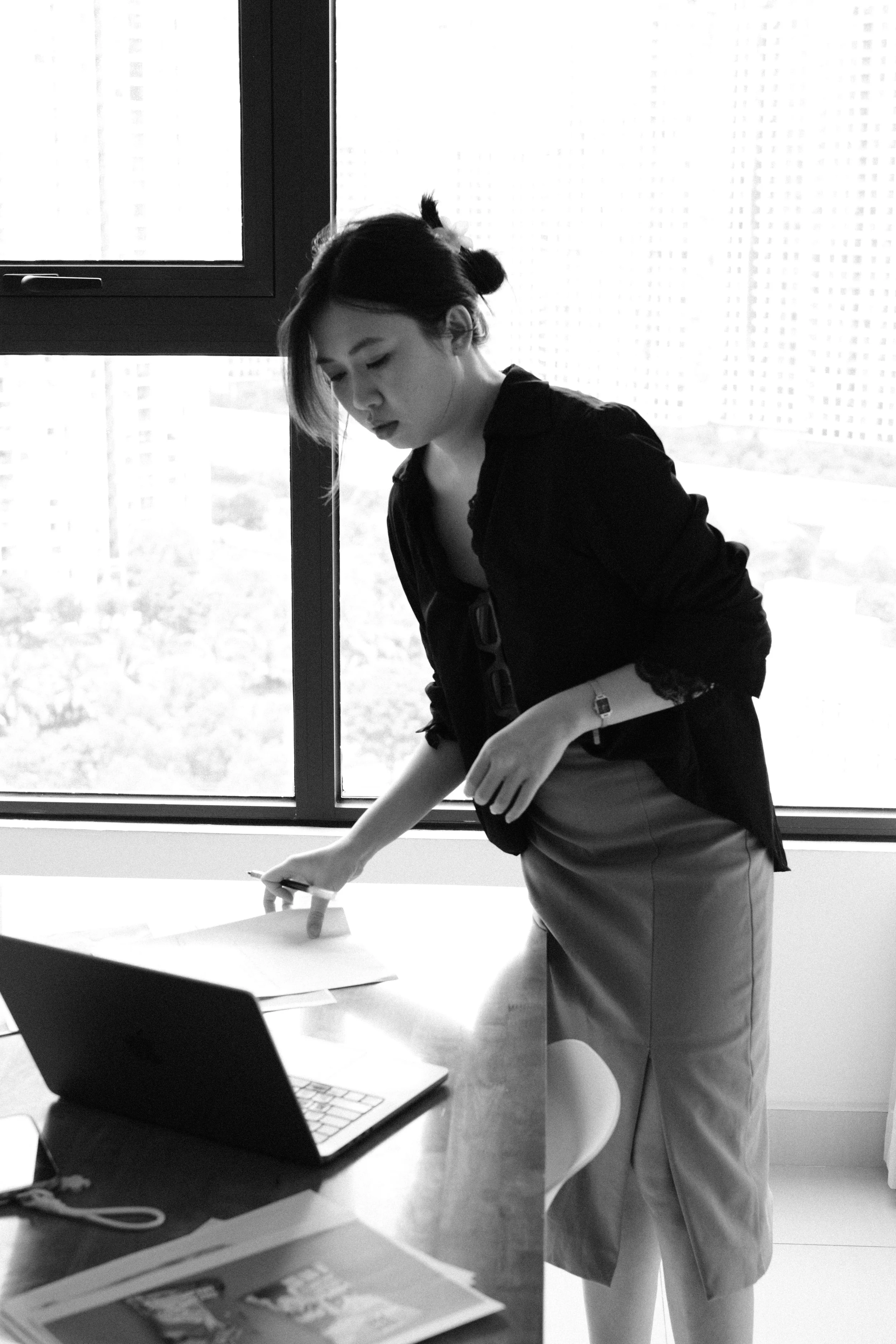 A woman dressed in business attire, with a bun hairstyle, is standing by a table, wiping it with a cloth. There is a laptop and some papers on the table. She is in an office near a large window.