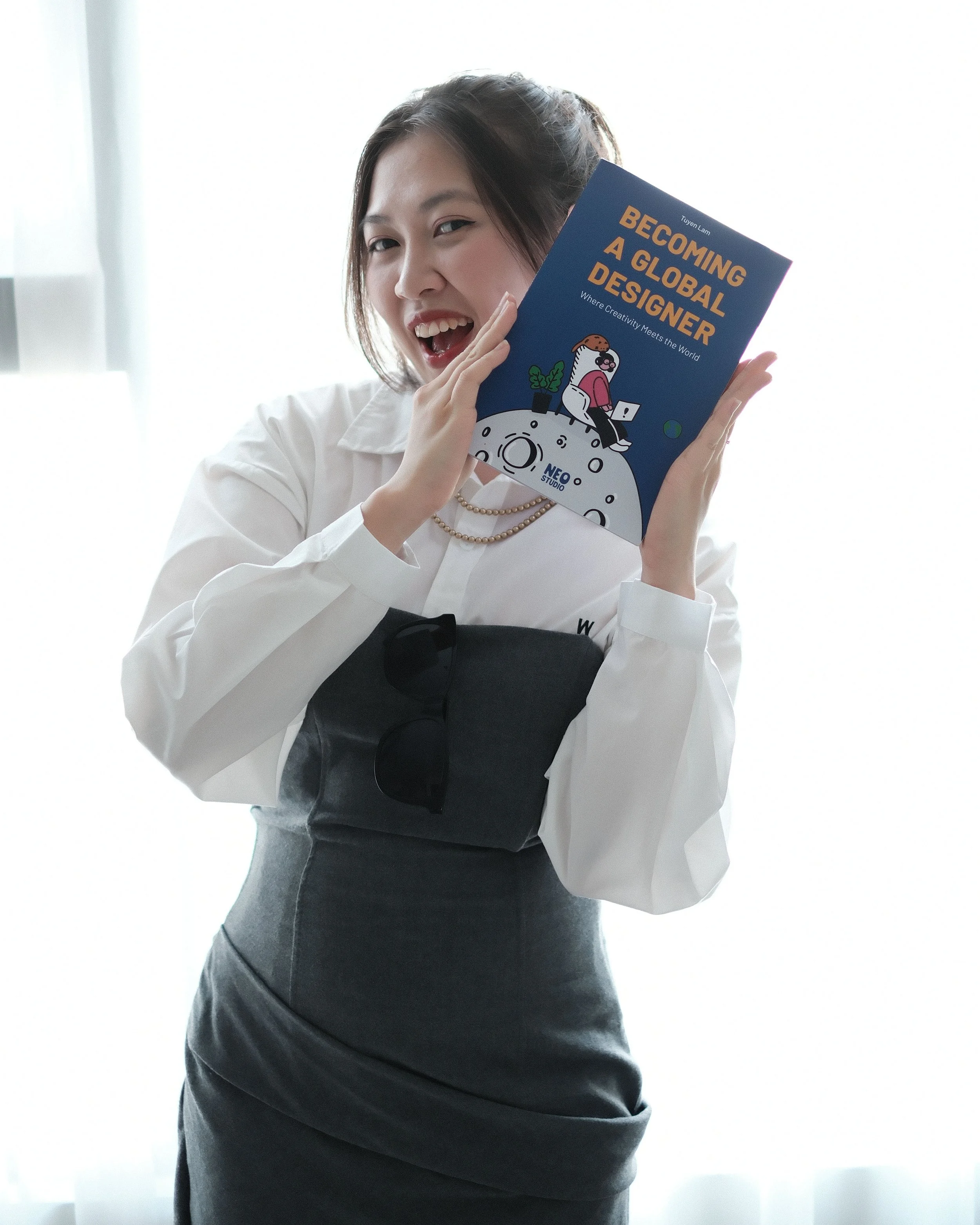 A woman dressed in business attire holding a book titled "Becoming a Global Designer" with a cartoon illustration of a woman sitting on a moon with planets and stars in the background, in front of a bright window.