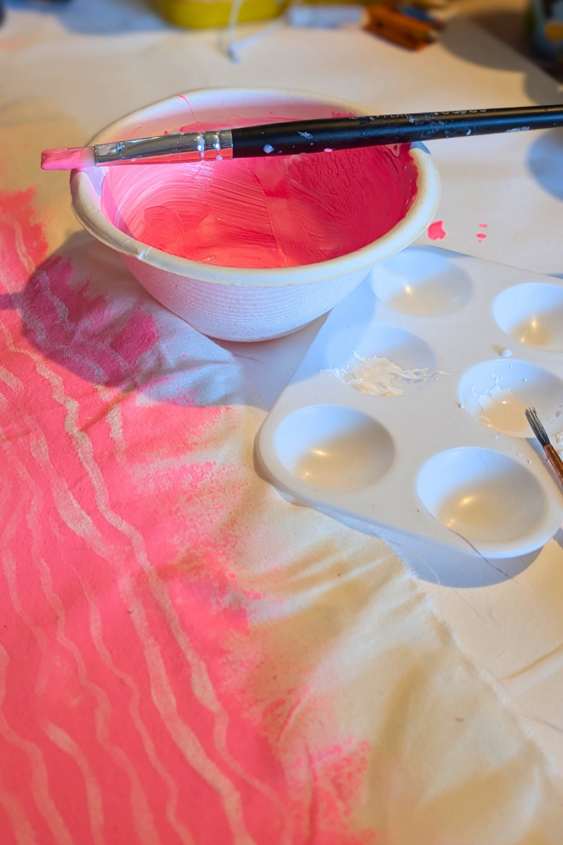 A white bowl of bright pink paint with a brush resting across it, sitting on fabric marked with soft pink wavy brushstrokes. A paint palette sits alongside.