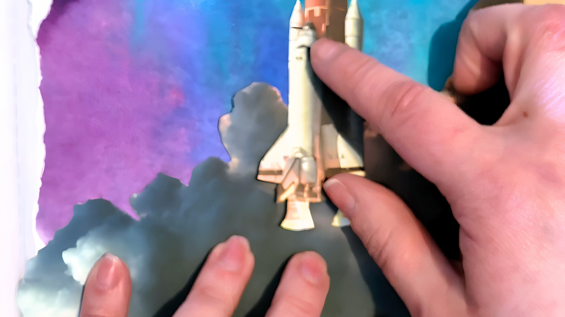 Hands arrange cutout images of clouds and a space shuttle on a blue and purple watercolour background.