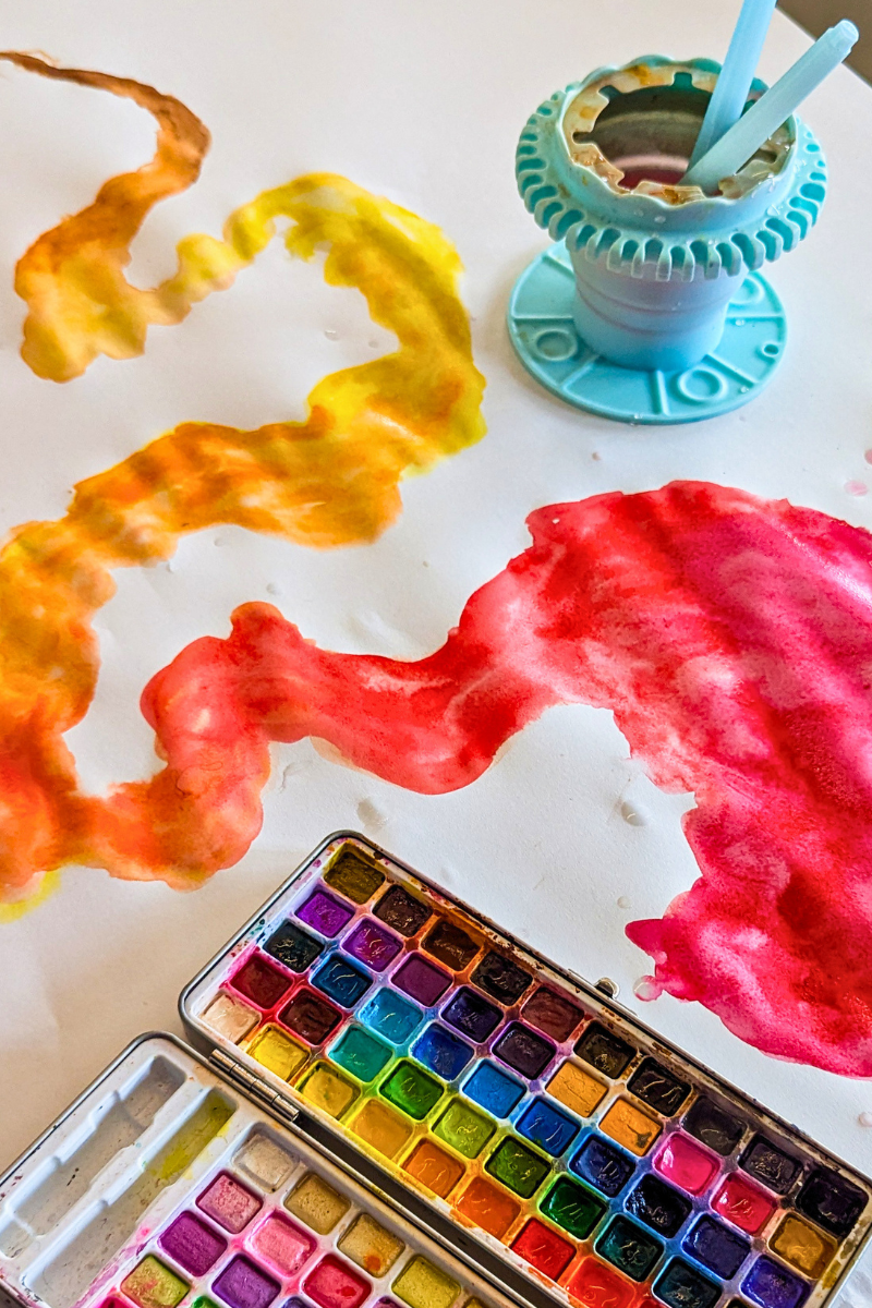 Loose swirls of yellow, orange and pink watercolour trailing across white paper, with an open tin of watercolour paints and a water pot nearby.