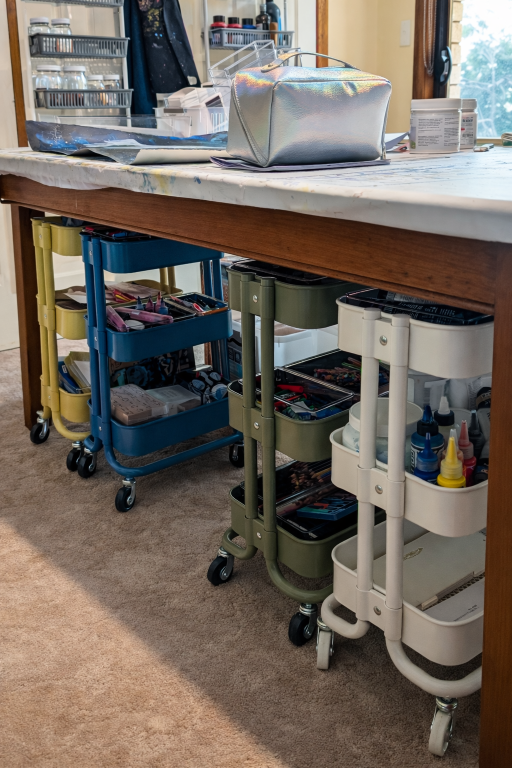 Rolling storage trolleys under a studio table holding art materials including pencils, paint bottles, and tools.
