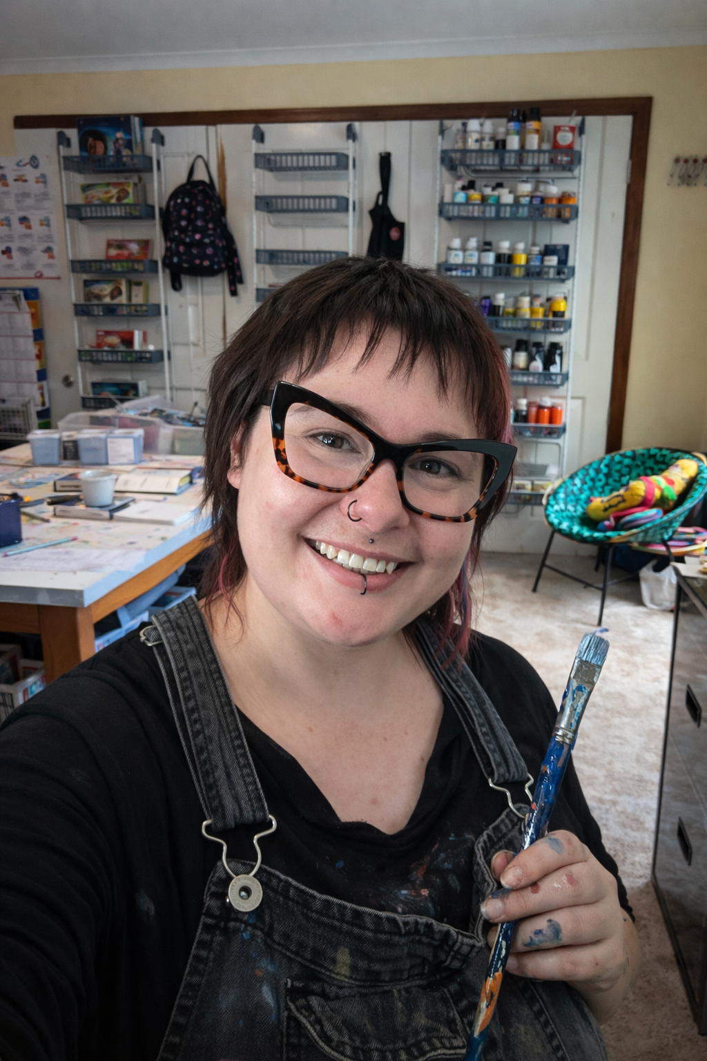 Tegan Bailey standing in her studio holding a paintbrush, with shelves of art materials behind her.