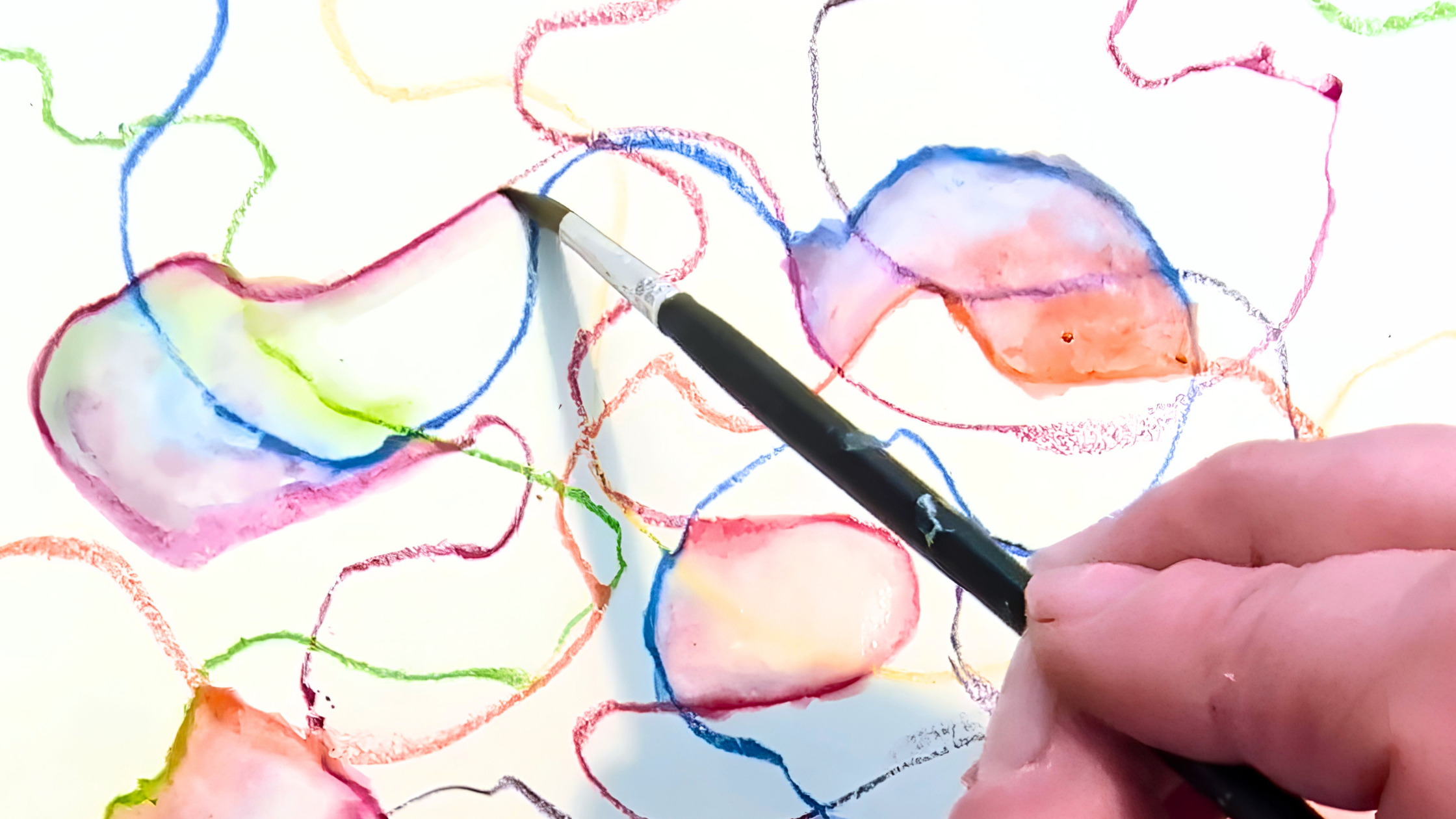 A quiet studio table seen from above — a hand holds a paintbrush as they work with watercolour pencil marks in reds, purples, blues and greens.