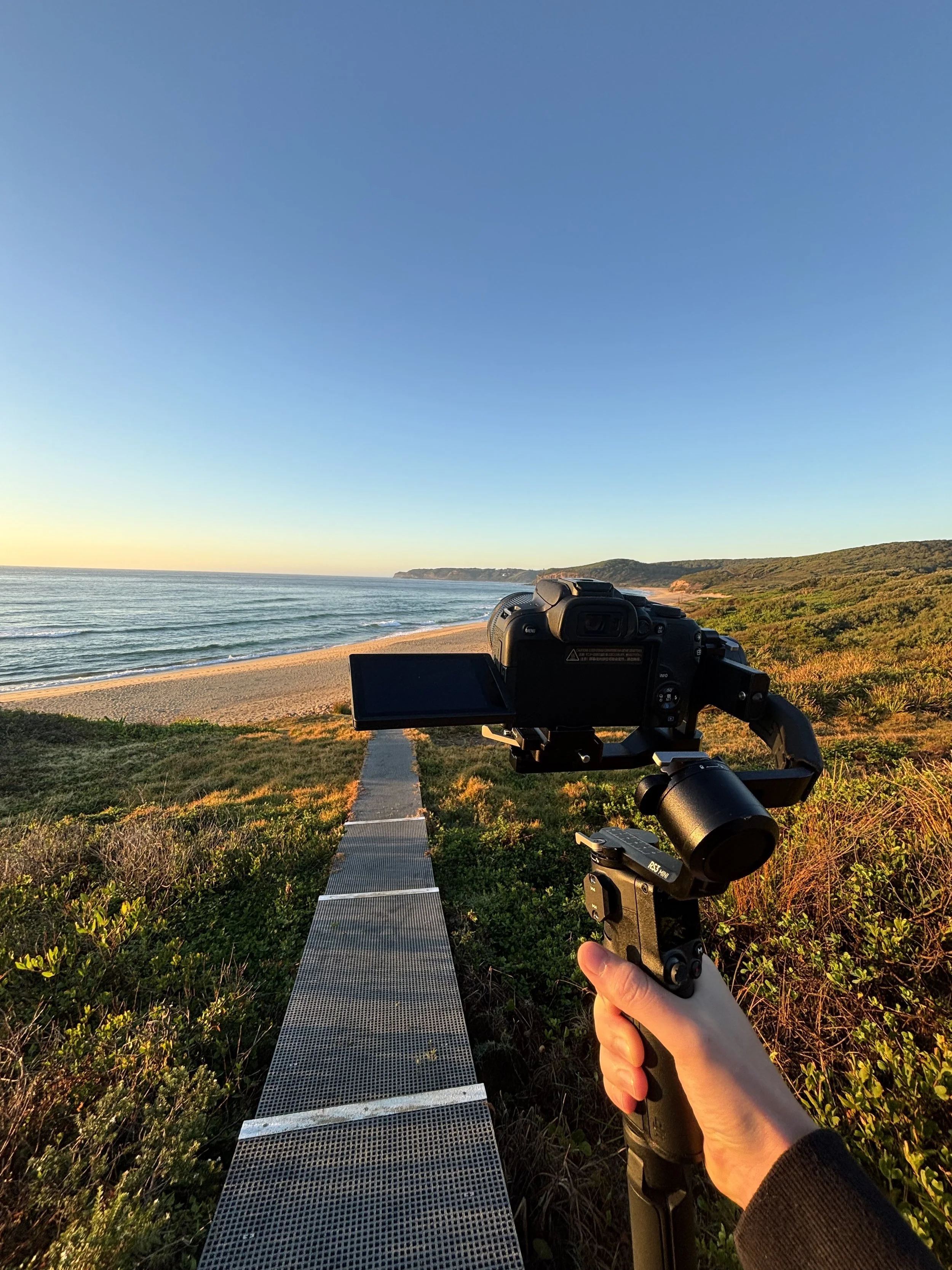 Person holding a camera on a stabilizer, aimed at the ocean and beach at sunset, with a walkway leading toward the shoreline and distant cliffs.