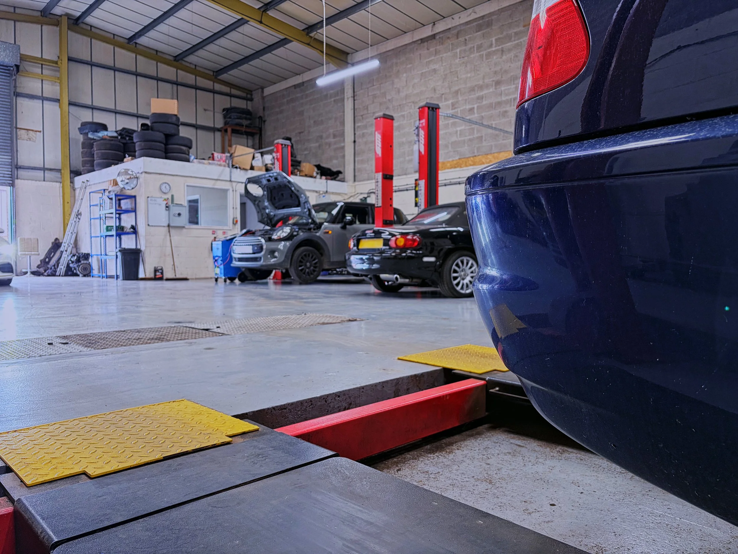 Inside a garage or auto repair shop, a car is on a hydraulic lift. There are tires stacked on a shelf, a worktable, and a small station with various tools and equipment. The environment has high ceilings and metal support beams.