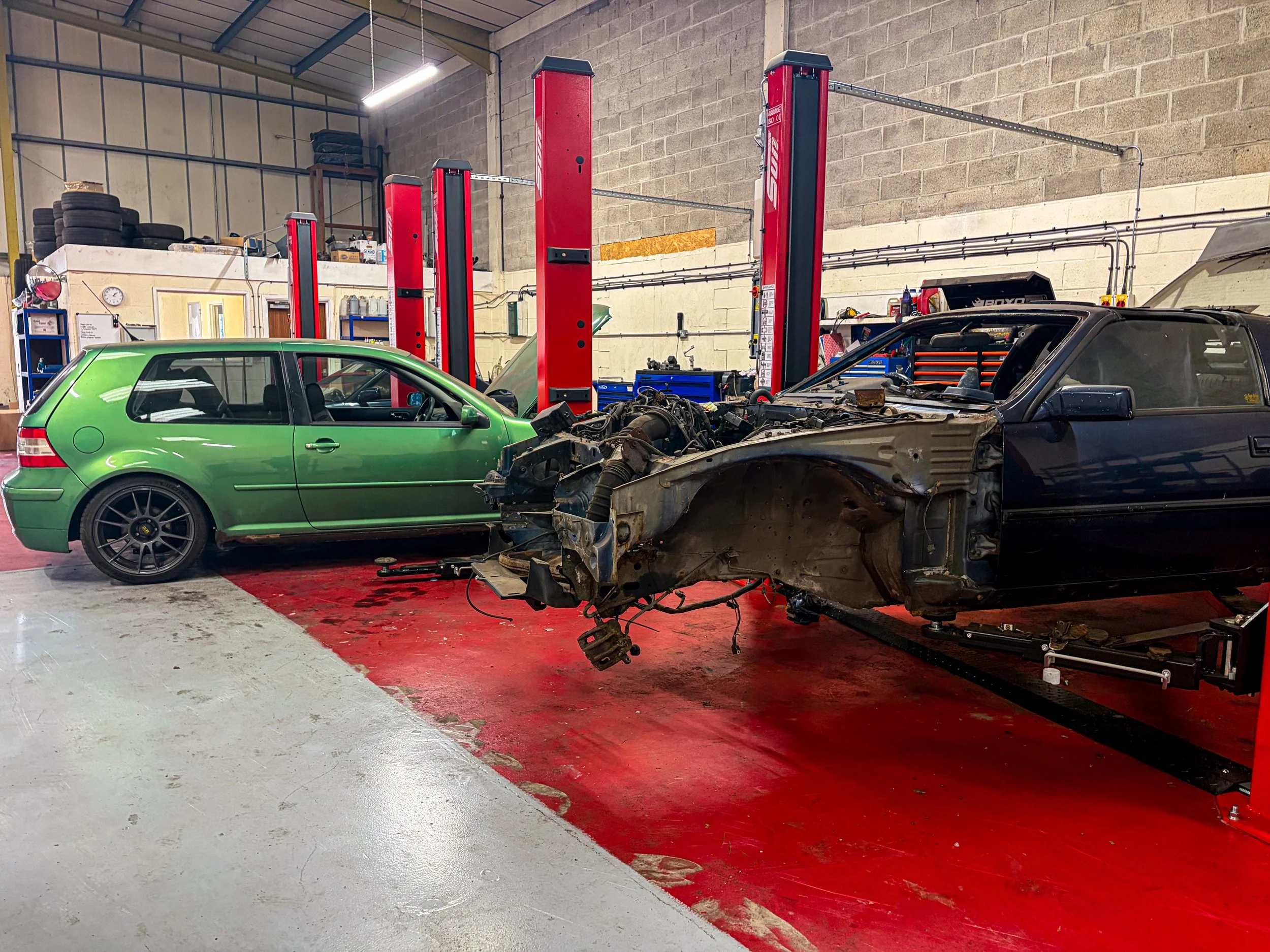 Inside an auto repair shop with two cars, one green and one black, undergoing repairs. The green car is intact, while the black car is partially disassembled, with its front end removed, exposing its engine and chassis, and is elevated on a hydraulic lift.