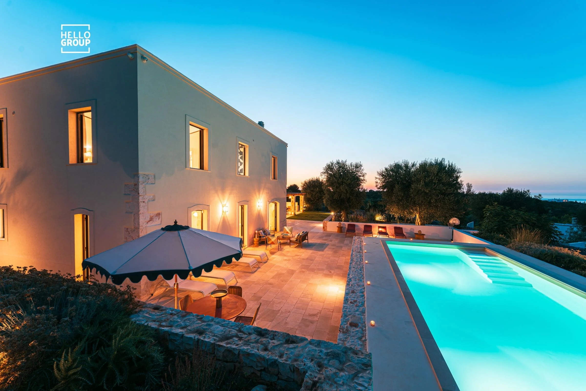 A modern house with outdoor patio lighting, patio furniture, a swimming pool with underwater lights, and trees in the background during sunset or twilight.