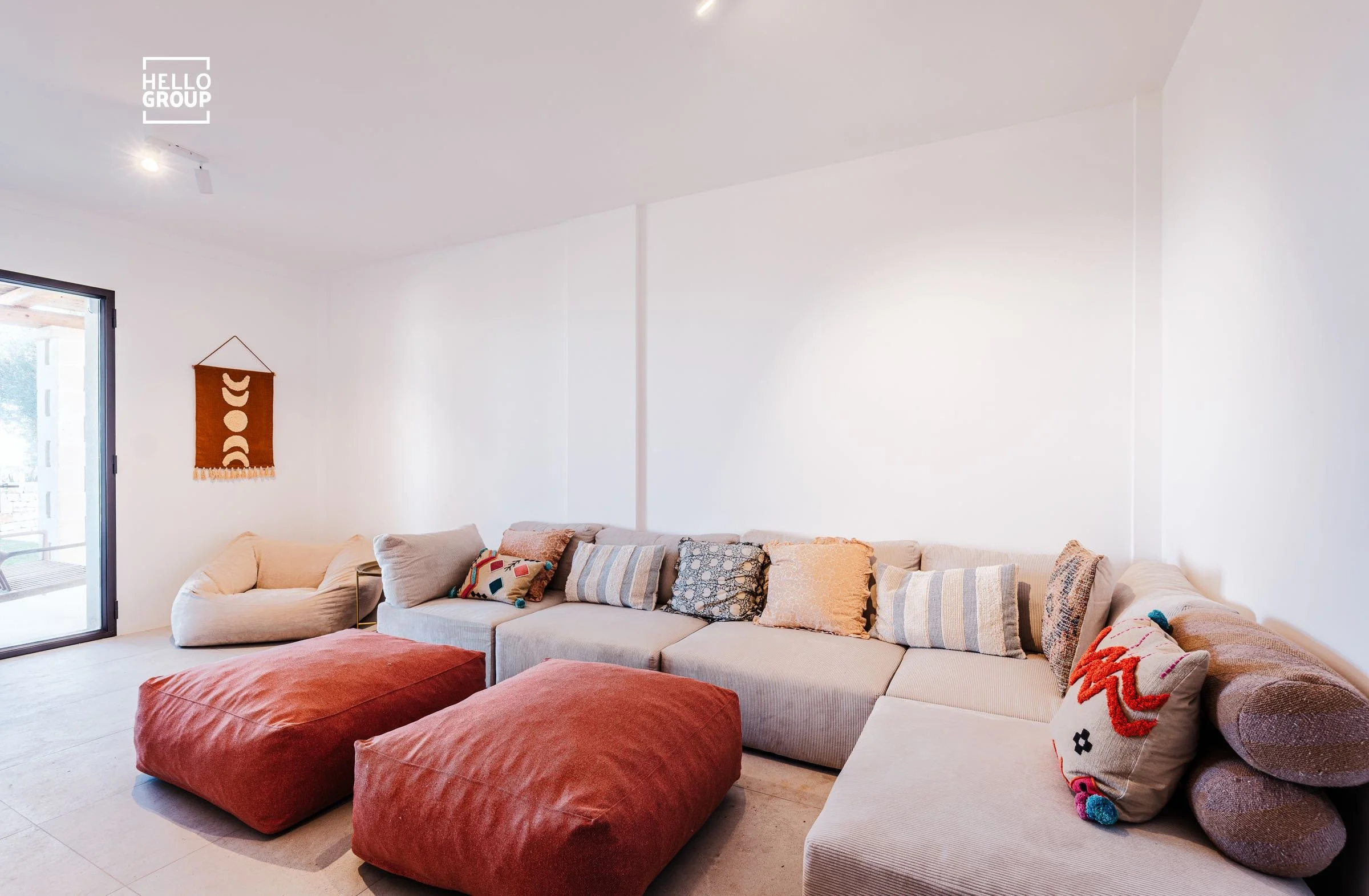 Living room with white walls and a large L-shaped beige sofa adorned with multiple patterned and textured cushions, two rust-colored ottomans in front, a beige armchair, and a wall hanging with a geometric design; sliding glass door on left leading outside.