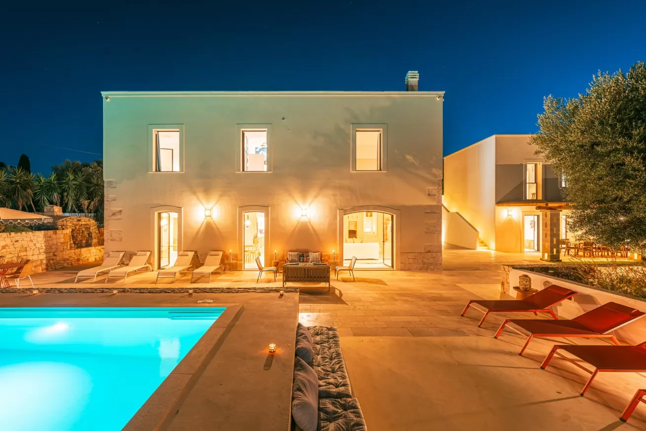 A modern house illuminated at night with a swimming pool in the foreground and outdoor seating areas on a stone patio.
