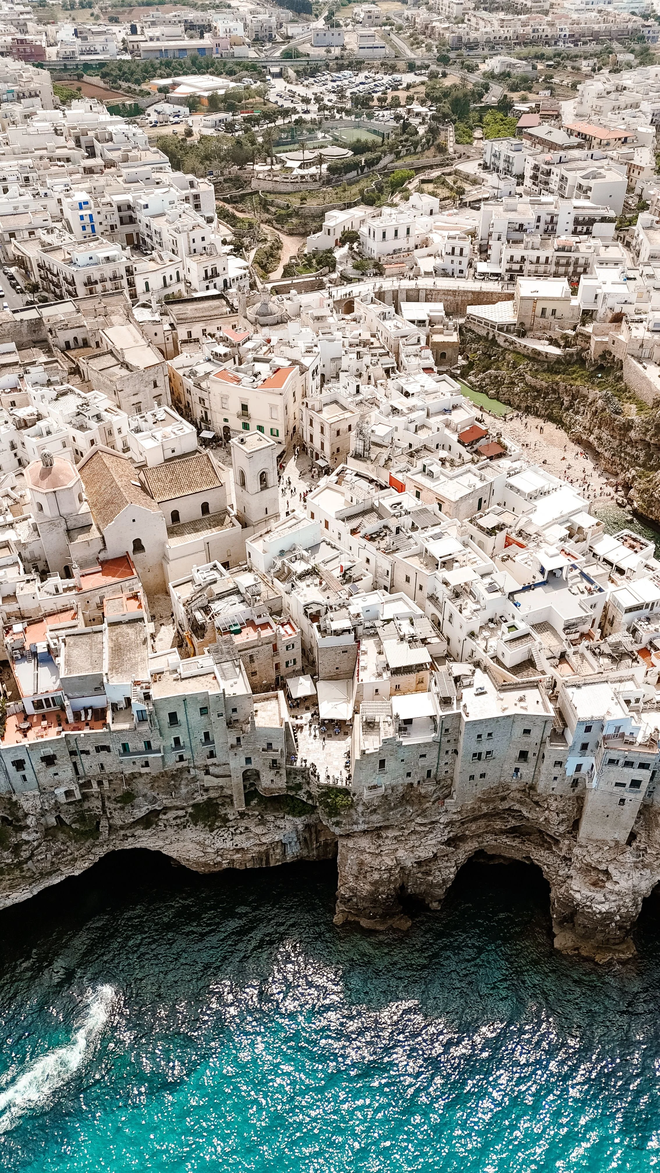Aerial view of a coastal city with white buildings, a church, and a rocky shoreline with caves, overlooking a blue sea.