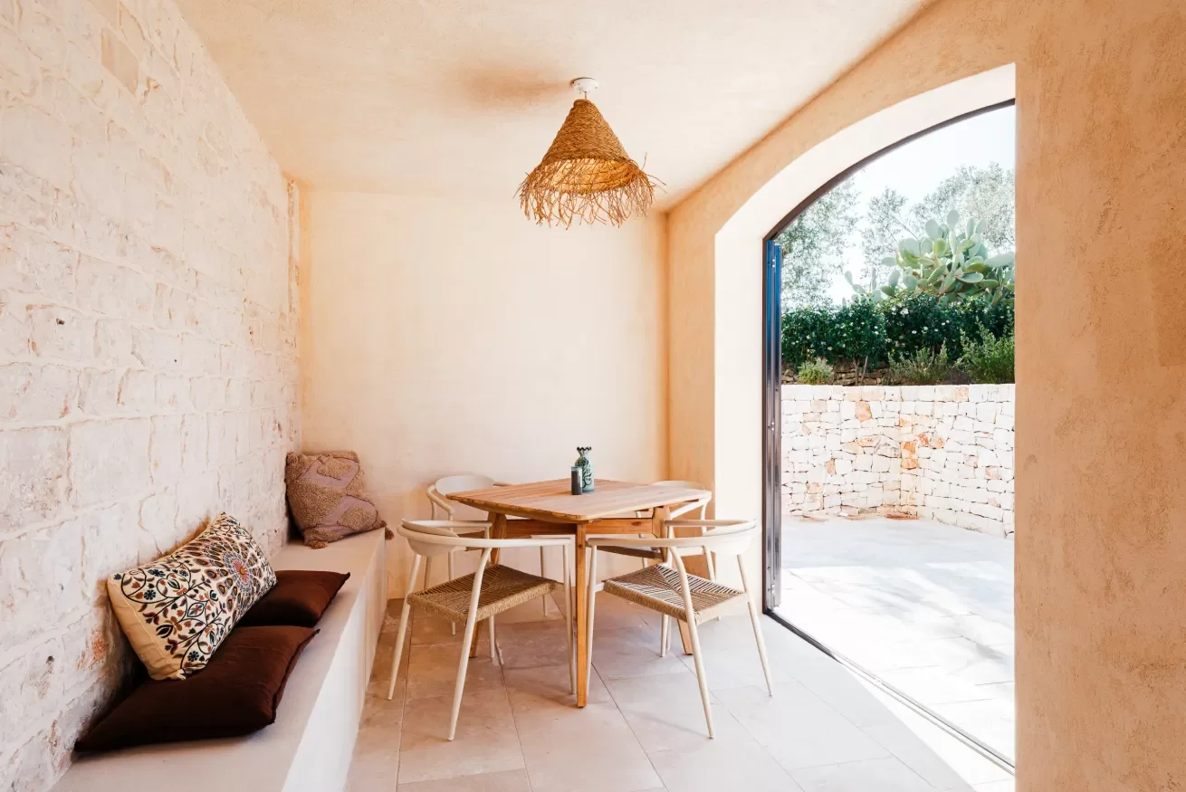 A cozy indoor dining area with a white brick wall, a built-in bench with decorative pillows, a round wooden table with four chairs, and a large arched outdoor entrance leading to a stone patio and garden with greenery.