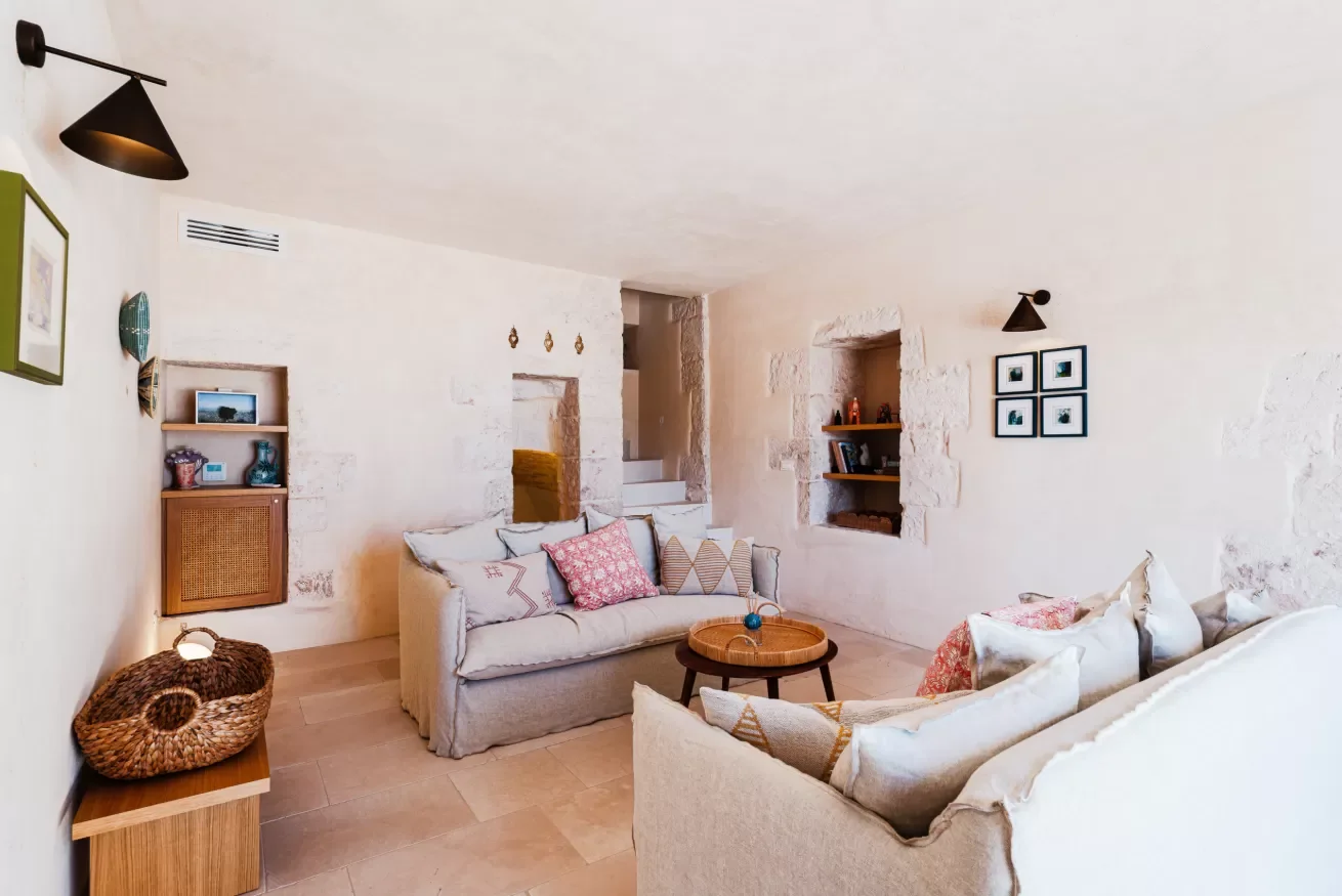 Cozy living room with white stone walls, beige sofas with patterned pillows, a round wooden coffee table, built-in wall shelves, framed artwork, and eclectic decor.