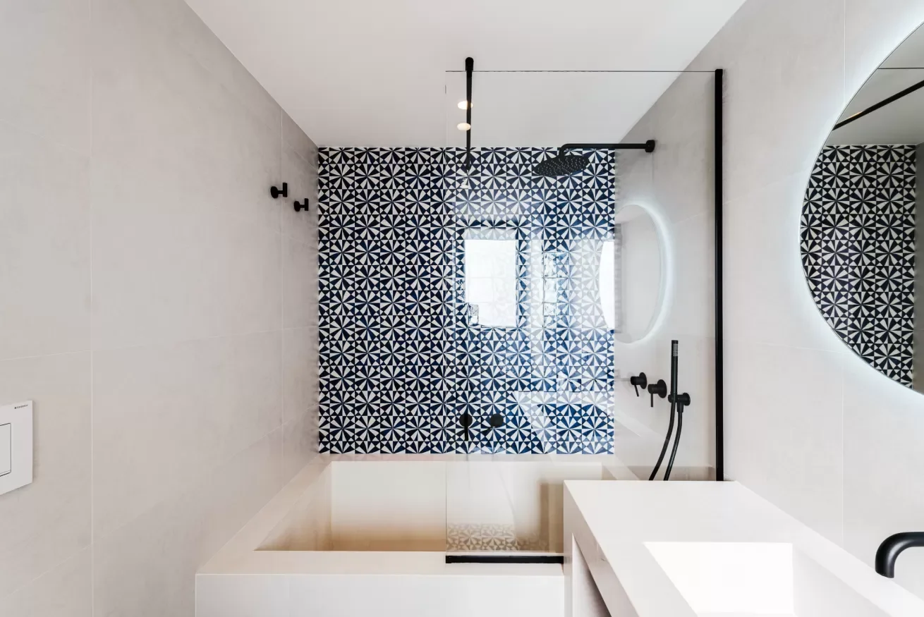 Modern bathroom with a walk-in shower featuring a glass door, black fixtures, and geometric black and white patterned tiles on the wall. A small window provides natural light, and a round mirror is visible on the right side.