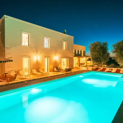 Night view of a lit swimming pool with lounge chairs and a modern building in the background.