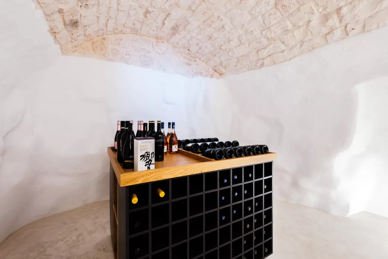 Wine bottles and boxes on a wooden table, with a wine rack below, in a room with white walls and a stone vaulted ceiling.