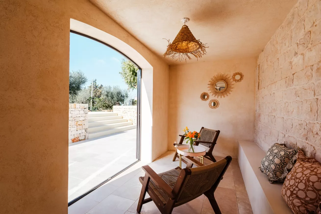 Cozy sitting area with two wooden chairs, a small round table with flowers, wall mirrors, and a large open doorway leading outside with steps and trees