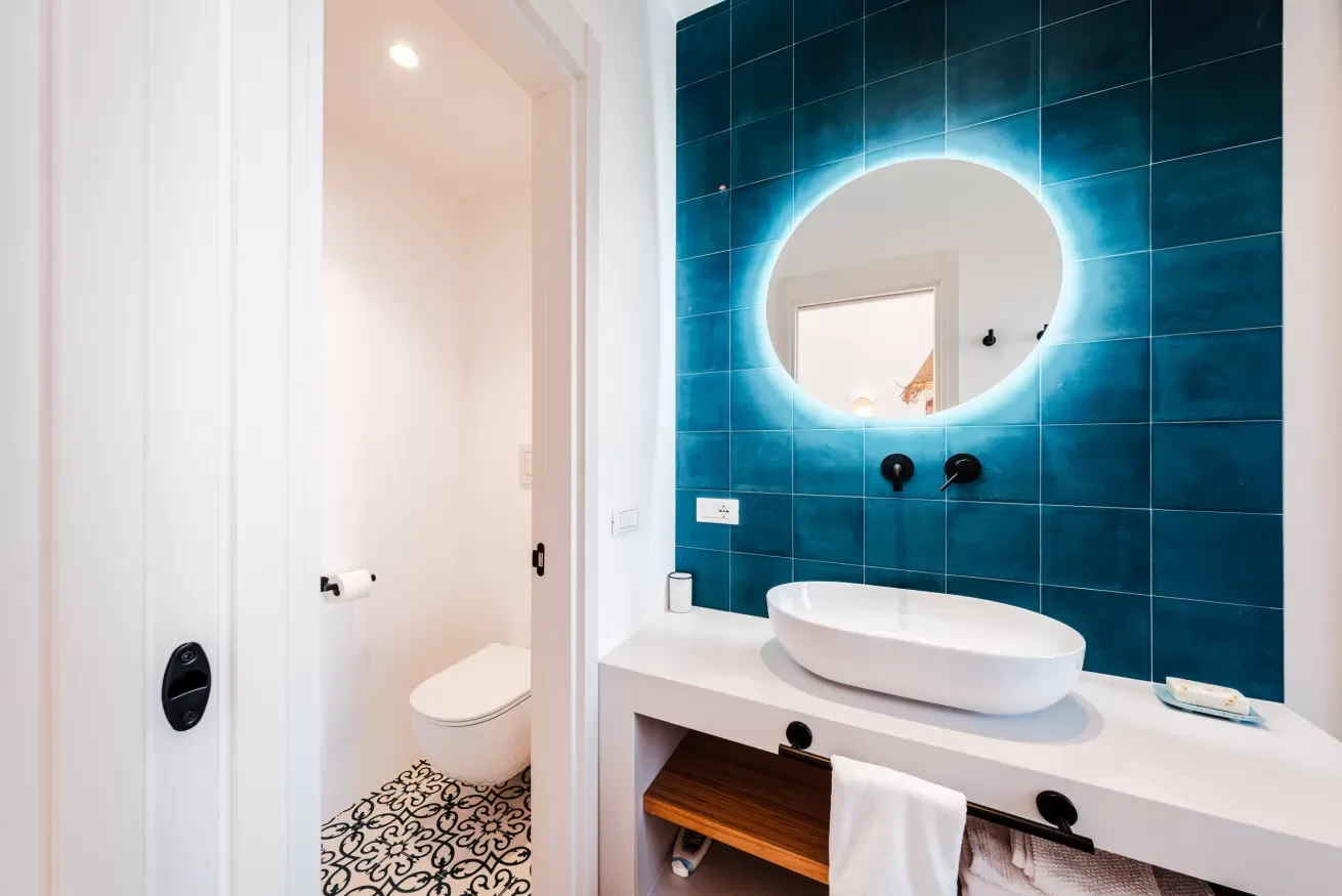 Modern bathroom with a vessel sink, large round backlit mirror, blue tiled wall, and a separate toilet area with patterned floor tiles.