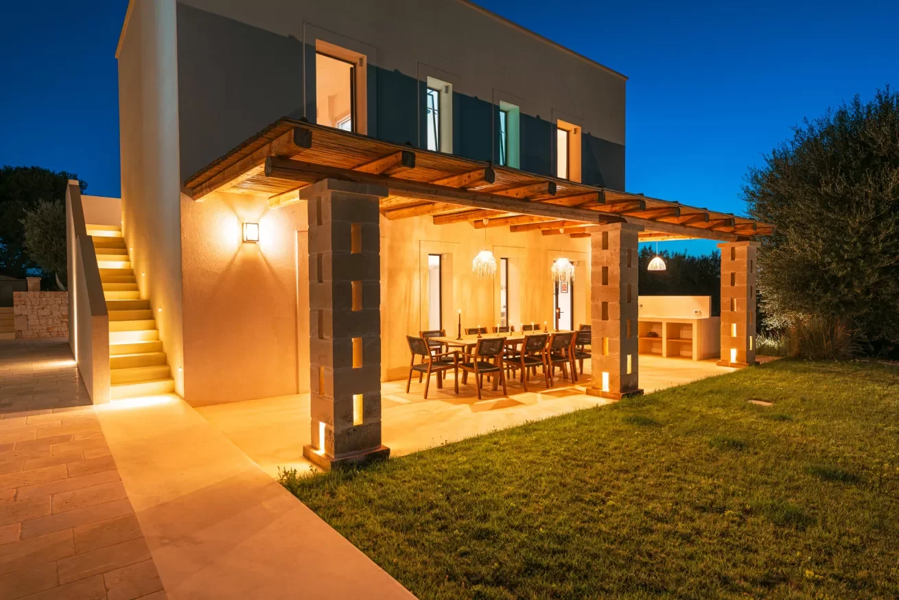 A modern house with an outdoor dining area illuminated at dusk, featuring a large wooden dining table and eight chairs under a wooden pergola, with stairs leading to the upper level on the left and a grassy yard to the right.