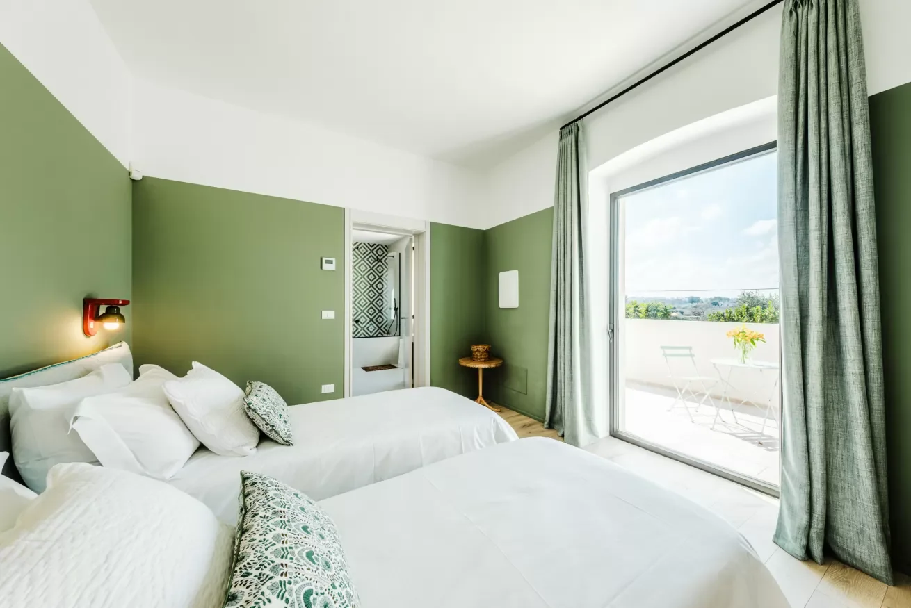 Bedroom with two white beds, green walls, a sliding glass door leading to a balcony, and outdoor furniture with a small table and flowers.