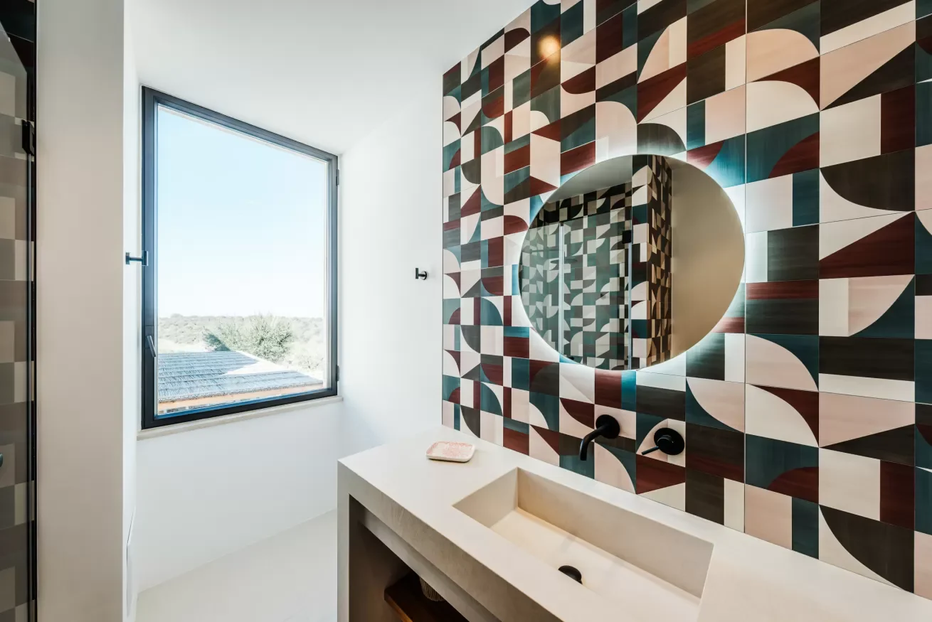 Modern bathroom with a large window, a geometric patterned tile wall, a backlit round mirror, and a concrete vanity with a built-in sink.