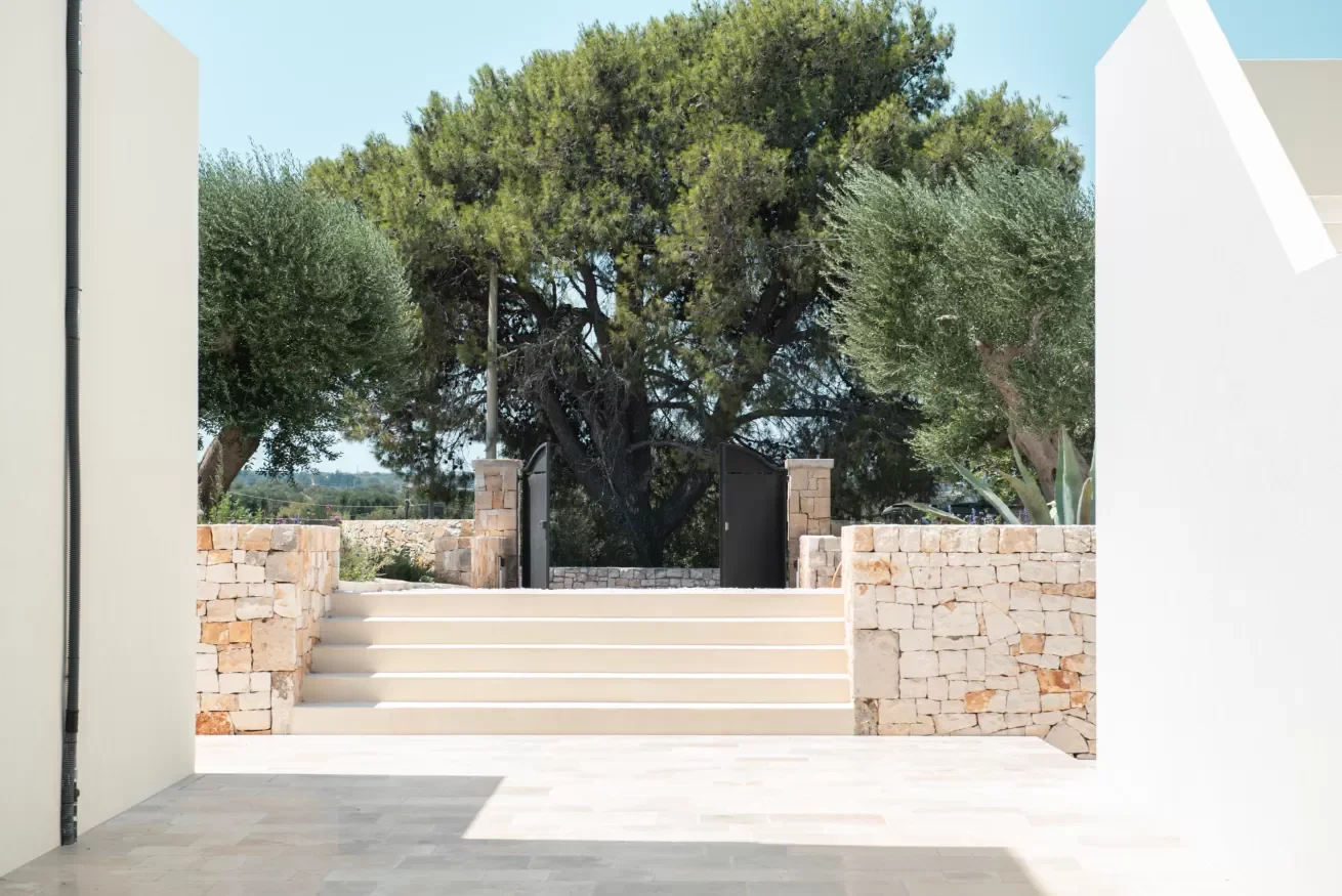 Outdoor area with stone steps leading to a black gate flanked by stone walls, with trees and greenery in the background, under a clear sky.