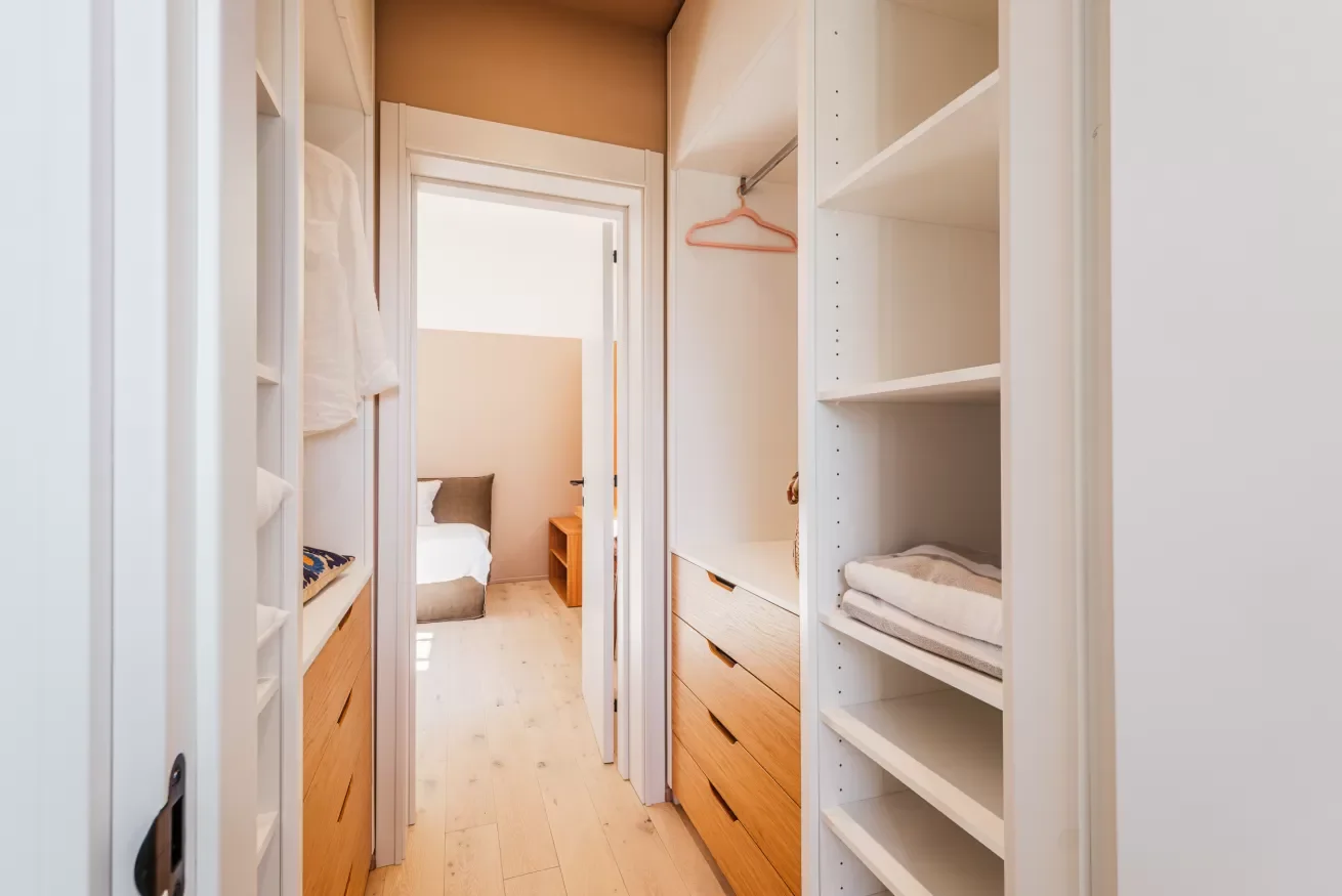 View of a walk-in closet with white shelving and drawers, leading to a bedroom with a bed and a nightstand.