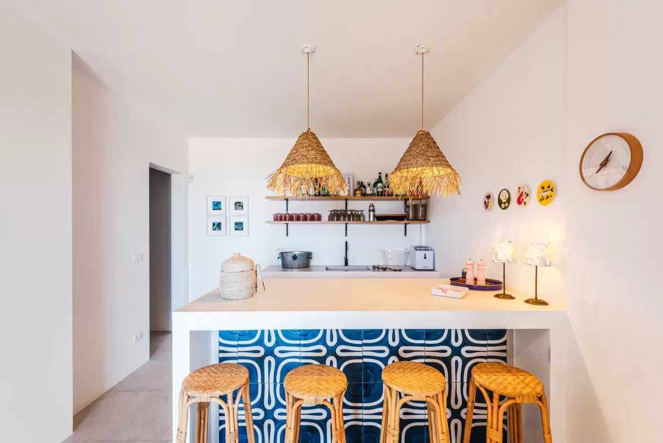 Modern kitchen with two woven pendant lights, open shelves with bottles and glasses, light-colored countertops, and four barstools with woven seats in front of a blue and white patterned panel. Decor includes small lamps, plates, and a wall clock.