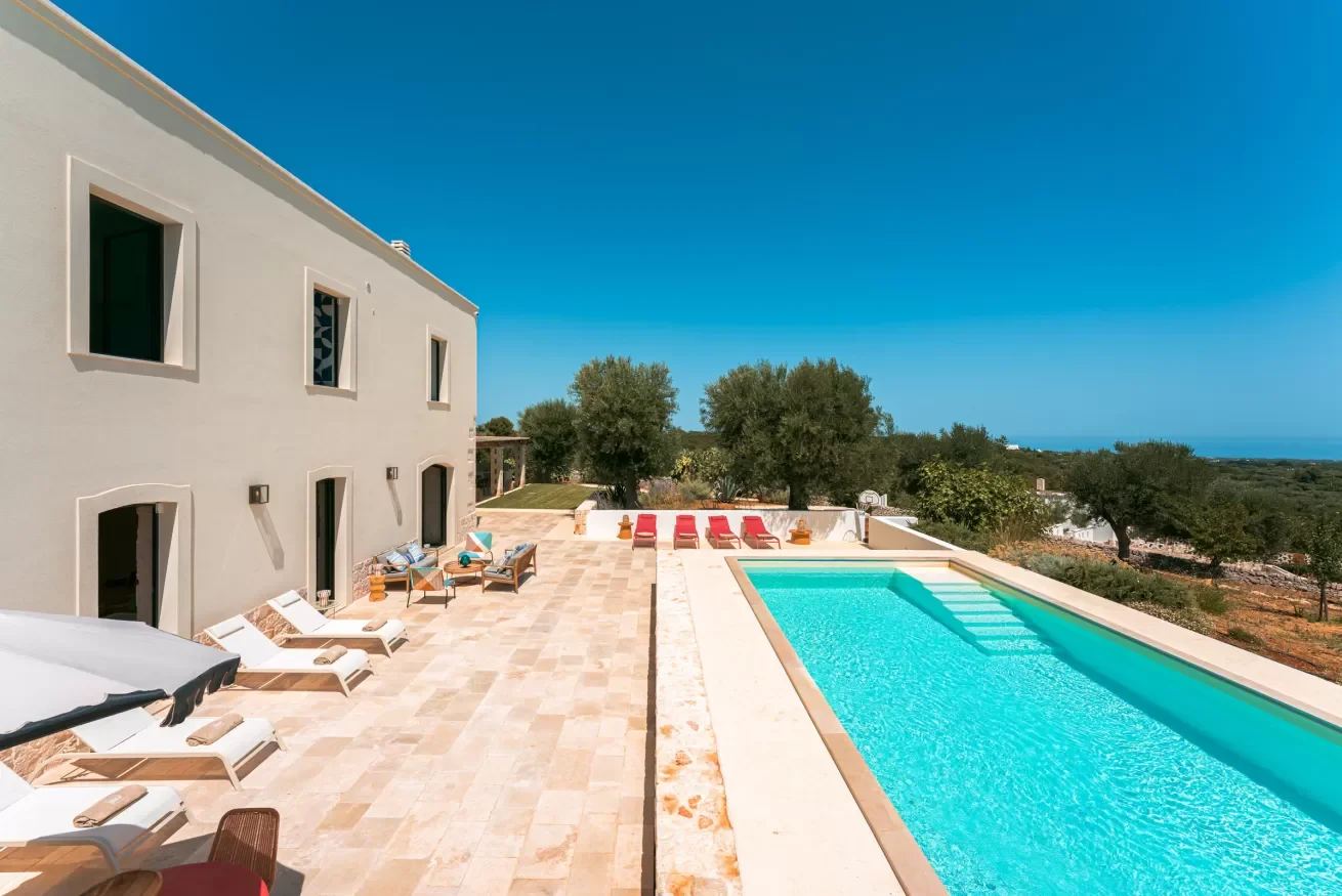 Outdoor pool area with lounge chairs and a building on the left, trees in the background, and a clear blue sky.