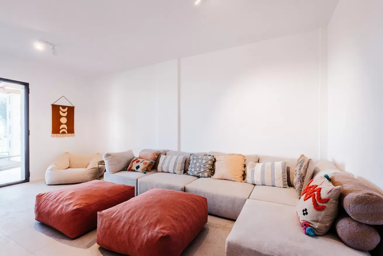 Living room with a long beige sectional sofa, various patterned pillows, two large rust-colored ottomans, a beige armchair, a wall hanging piece, and an open sliding glass door leading outside.