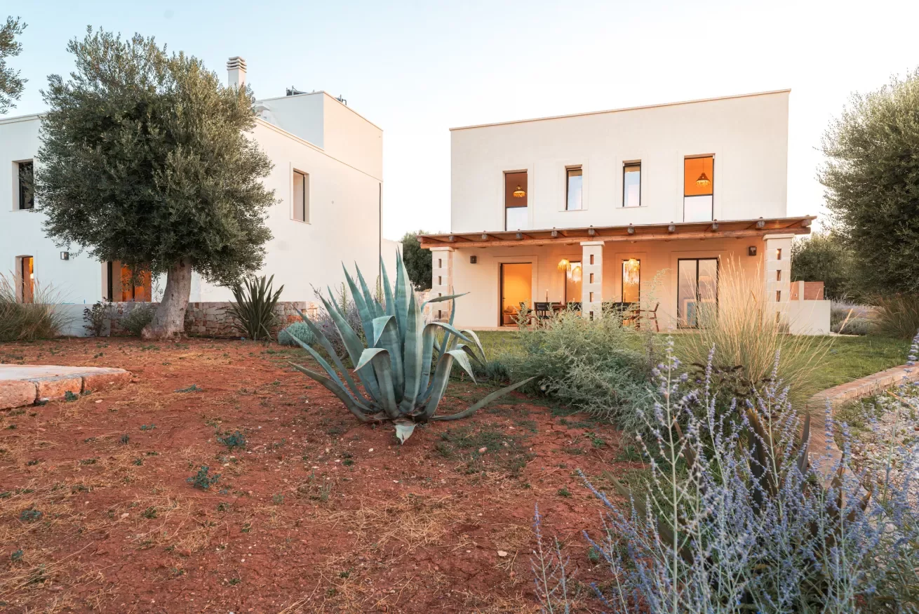 Modern two-story house with a white exterior, large windows, and a wooden porch. The front yard has desert plants, including an agave, grasses, and a tree, with reddish soil and a stone pathway.