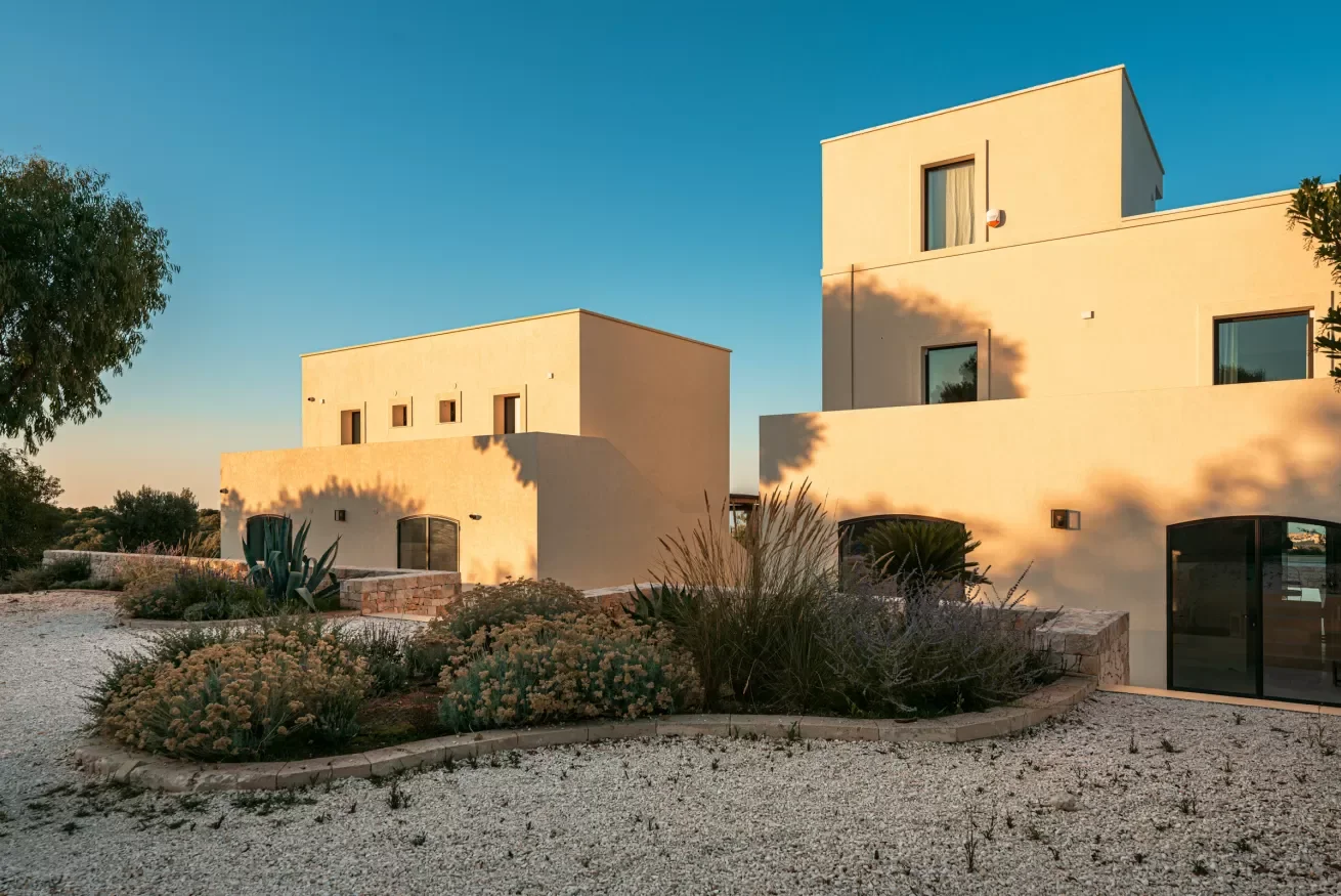 Modern beige residential buildings with flat roofs, surrounded by desert landscaping, with shadows cast by trees during sunset.
