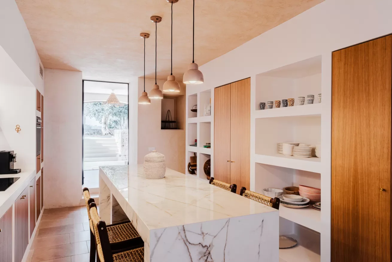Modern kitchen with a marble island, wooden cabinetry, open shelves with dishes and cups, and hanging pendant lights.