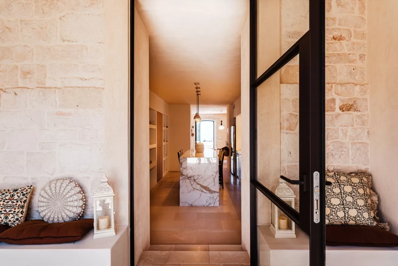 A view through a glass door into a modern, warmly lit kitchen and dining area with a marble island, dining table, and chairs, featuring a neutral color palette and decorative pillows on built-in seating.