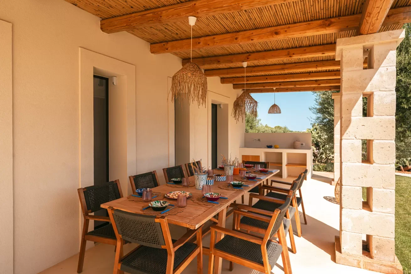 Outdoor dining area with a wooden table set with dishes and cups, surrounded by black chairs, under a wooden ceiling with hanging woven light fixtures, and a partial stone wall in the background.