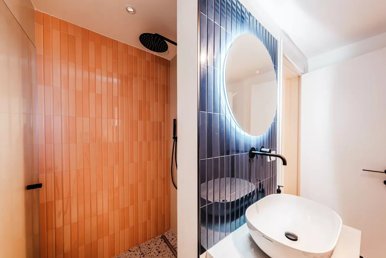 Bathroom with a shower area on the left with orange tiles and a rain shower head, and a sink with a large round mirror and blue tiled wall on the right.