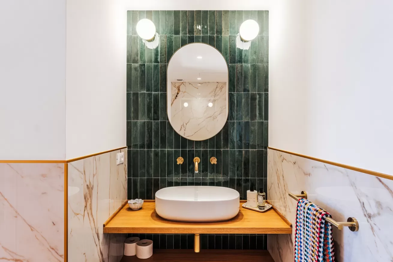 Modern bathroom with an oval mirror, green tiled wall, white vessel sink, stone wall accents, and gold fixtures.